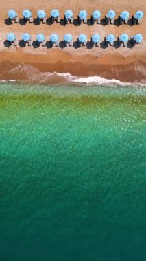 Mavi güneş şemsiyeleri ve turkuaz deniz suyuyla cennet gibi kumlu sahilin yukarıdan görünüşü. Dikey video.