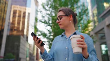Gözlüklü beyaz kadın sokakta duruyor, akıllı telefon kullanıyor ve kahve içiyor. Yörünge atışı. Arka planda gökdelenler var. İletişim, iş günü, yoğun yaşam konsepti.
