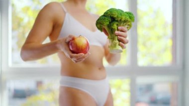 Bir kadın elinde brokoli ve elmayla dans ediyor. Hangisinin daha iyi olduğuna o karar verir. Sağlıklı beslenme kavramı.
