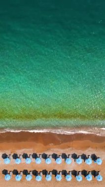 Mavi güneş şemsiyeleri ve turkuaz deniz suyuyla cennet gibi kumlu sahilin yukarıdan görünüşü. Dikey video.