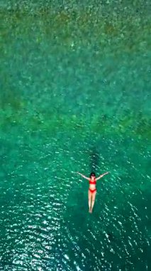 Kırmızı mayo giymiş bir kadının hava görüntüsü, kristal berrak deniz sularında rahatlama somutlaştırıcı bir şekilde sırtüstü uzanır. Yaz tatilleri. Dikey video