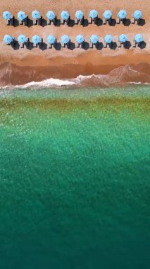 Mavi güneş şemsiyeleri ve turkuaz deniz suyuyla cennet gibi kumlu sahilin yukarıdan görünüşü. Dikey video.