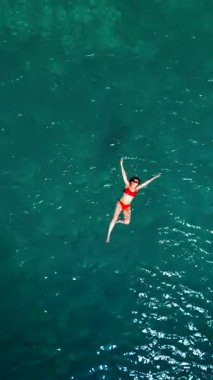 Kırmızı mayo giymiş bir kadının havadan görünüşü, kristal berrak deniz sularında rahatlama somutlaştırıcı bir şekilde zahmetsizce sırt üstü yüzüyor. Yaz tatilleri. Dikey video