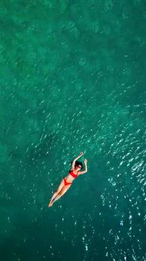 Kırmızı mayo giymiş bir kadının hava görüntüsü, kristal berrak deniz sularında rahatlama somutlaştırıcı bir şekilde sırtüstü uzanır. Yaz tatilleri. Dikey video