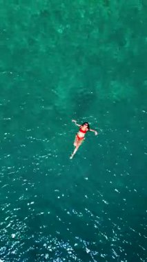 Kırmızı mayo giymiş bir kadının havadan görünüşü, kristal berrak deniz sularında rahatlama somutlaştırıcı bir şekilde zahmetsizce sırt üstü yüzüyor. Yaz tatilleri. Dikey video