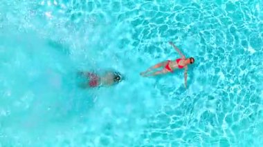 Çift havuzda rahatlar. Bir erkek havuza atlarken ve bir kadın sırt üstü yatarken yüzerken yukarıdan aşağıya doğru bak. Yavaş çekim