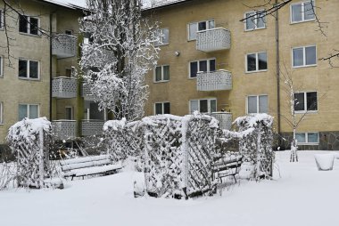 Ağaçlı bir yerleşim yeri ve balkonlu sarı bir apartmanın önünde karla kaplı bir bank.