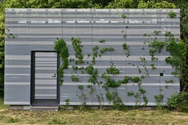 Metal kaplamalı modern bir yapı kısmen yeşil tırmanma bitkileriyle kaplı. Binanın merkezi bir kapısı var ve yemyeşil ağaçlar ve yemyeşil bir zemin üzerine kurulmuş. Yapının önündeki zemin çimenli, katkıda bulunan 