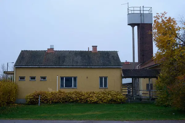 stock image A simple, yellow single-story building with a tile roof and small, rectangular windows, set against an overcast sky. The building is flanked by autumnal shrubs with yellow leaves and a grassy lawn. In the background, a tall, brick water tower with a 