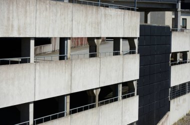 Bu görüntü, çok katlı bir otoparkın dış görünüşünü gösteriyor. Beton yapı, kentsel mimarinin tipik ham, endüstriyel tasarımını sergiliyor. Binanın birden fazla katı görülebiliyor. Beton duvarlar arasında açık alanlar var.