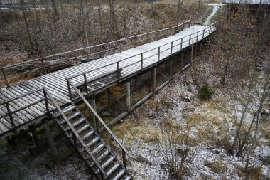 Bu resim, kış manzarasında süzülen hafif kar tozlarıyla kaplı yükseltilmiş bir tahta kaldırımı gösteriyor. Patika, merdivenler ve tırabzanlarla tamamlandı, ormanlık bir alanda ilerliyor, kış yürüyüşü ve doğa için manzaralı bir rota oluşturuyor.