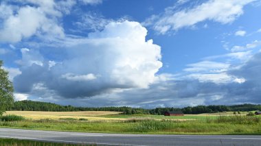 Büyük, kabarık bulutlarla dolu dramatik bir gökyüzü altında geniş alanlar barındıran pitoresk bir kırsal alan. Yeşillik ve çayırların ufka doğru uzandığı bir yol ön planda uzanıyor. Sahnede birkaç ahır ve yama var.