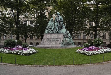 Fotoğrafta, Fin ulusal destanı Kalevala 'nın yazarı Elias Lonnrot' un ayrıntılı bronz heykeli yer alıyor. Bereketli yeşil ağaçlar ve renkli çiçekliklerle dolu sakin bir parkta yer alan anıtta, bir kitapla oturmuş Lnnrot, iki figür yer alıyor.
