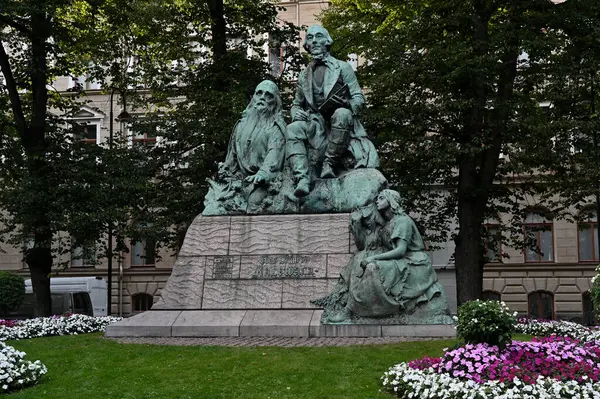 stock image The photo features a detailed bronze statue of Elias Lonnrot, the author of the Finnish national epic Kalevala. The monument, set in a serene park with lush green trees and colorful flowerbeds, depicts Lnnrot seated with a book, while two figures 