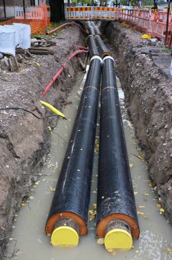 The image showcases a construction site focused on installing large underground pipes. Two large insulated pipes are visible in a trench filled with water, likely part of a district heating or water distribution system. The trench is surrounded by co clipart