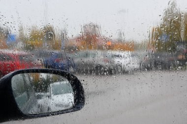 This photo captures a rainy day scene from inside a car, where raindrops have accumulated on the windshield, blurring the view of the outside. The side mirror reflects part of the rain-splattered exterior, while in the background, parked cars and aut clipart