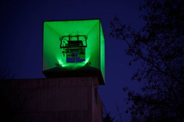 bell tower of the Lutheran Evangelical Church in Jarvenpaa, Finland with green illumination at night against a deep blue evening sky. The minimalist architectural design of the structure is framed by silhouettes of tree branches, adding an organic clipart