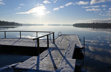 Açık bir kış sabahında huzurlu bir göl manzarası. Karla kaplı bir ahşap iskele sakin, yansıtıcı suya doğru uzanıyor ve sakin ve sakin bir atmosfer yaratıyor. Güneş ışıl ışıl parlıyor, uzun gölgeler bırakıyor ve manzarayı yumuşakça aydınlatıyor.