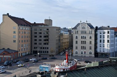 Helsinki 'de tarihi ve modern mimarinin bir karışımını sergileyen büyüleyici bir şehir manzarası. Ön planda, kafeterya ya ya da eğlence olarak yeniden tasarlanmış bir kırmızı ışık gemisi yer alıyor ve bir deniz dokunuşu ekleniyor. Geminin etrafı iyi korunmuş binalarla çevrili. 