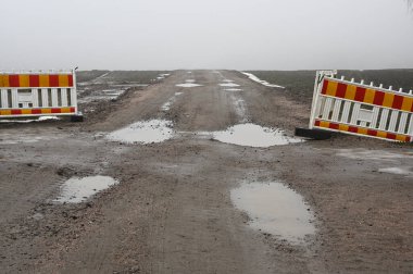 Çamurlu, asfaltsız bir yol sisli mesafeye kadar uzanıyor. Kırık inşaat bariyerleri kısmen engelliyor. Su birikintileri bulutlu gökyüzünü yansıtıyor, engebeli ve ıssız araziyi vurguluyor. Sahne, bitmemiş bir terk edilmişlik hissi veriyor. 