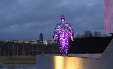 A striking contemporary sculpture illuminated with purple light, set against the backdrop of Helsinki's skyline on a winter evening. The reflective metallic figure exudes futuristic vibes, blending art with the Nordic city's urban and cultural landsc clipart