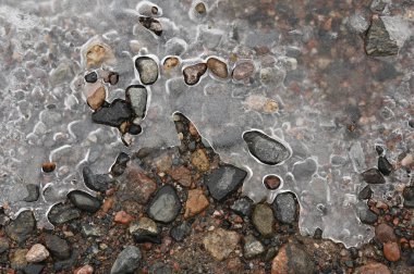 A close-up view of ice partially covering a rocky ground, capturing the intricate patterns formed as the ice begins to melt. The frozen surface reveals a mix of earthy-toned stones and pebbles, creating a beautiful contrast between solid and liquid s clipart