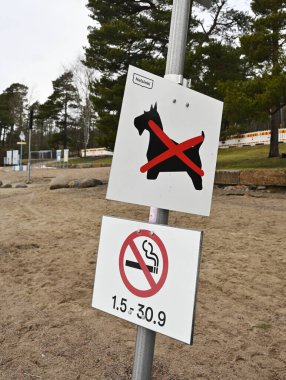 Finlandiya 'nın Helsinki kentindeki kumlu bir plaj bölgesinde, biri köpeklerin sigara içmesine izin verilmediğini gösteren iki tabela, diğeri ise 1-30 Eylül tarihleri arasında sigara içilmesini yasaklayan bir tabela bulunuyor. Arka planda p ile doğal bir kıyı ortamı vardır.