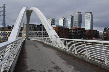 Finlandiya 'nın başkenti Helsinki' de bulunan modern beyaz kemerli bir köprü, sakin bir nehrin üzerinde uzanır ve su yüzeyindeki yapısını yansıtır. Arka planda, kentlerin vurgulandığı, yükselen konut binaları ve modern apartman kompleksleri yer alıyor. 