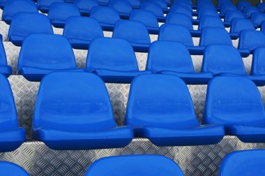 Metalik, desenli bir zeminde simetrik sırada dizilmiş boş mavi plastik stadyum koltuklarının yakın plan fotoğrafı. Canlı mavi renk gümüş metalle çelişiyor, görsel olarak çarpıcı ve tekrarlayan bir geometrik desen yaratıyor. Resim kapağı