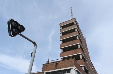 Minimalist modernist mimarisi olan kırmızı tuğlalı yüksek katlı bir binanın çarpıcı perspektifi ön planda benzersiz bir üçgen trafik ışığıyla yan yana. Sahne, açık mavi bir gökyüzüne göre ayarlanmış, geometrik çizgileri vurguluyor.