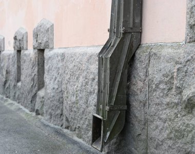 Pembe alçı kaplama duvarlı ve sağlam granit tabanlı bir binaya bağlı eski metal yağmur oluğunun yakın çekim görüntüsü. Yaşlandıkça kararan drenaj borusu kaldırımlı bir sokak seviyesinin hemen üstünde bitiyor.
