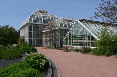 Helsinki Kaisaniemi Botanik Bahçesi 'ndeki güzel ve tarihi cam ev parlak mavi gökyüzü altında zarif Victorian tarzı mimarisini sergiliyor. Etrafı yemyeşil çiçeklerle, çiçek açan yataklarla ve düzenli çakıl yollarıyla çevrili manzara coşuyor.