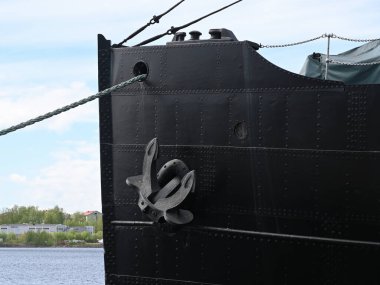 the imposing bow of a large black ship moored at a harbor, featuring a prominent steel anchor attached to its riveted metal hull. The industrial design and heavy-duty construction highlight the strength and functionality of maritime engineering. Visi