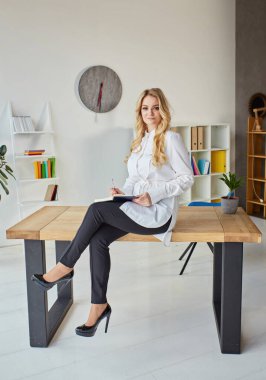 A blonde woman in a white shirt sits on a wooden office table, a notebook and a pen in her hands, makes notes while working at home