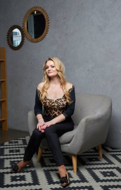 Portrait of beautiful blonde woman in black suit sitting on in classic interior. Fashion girl with makeup.