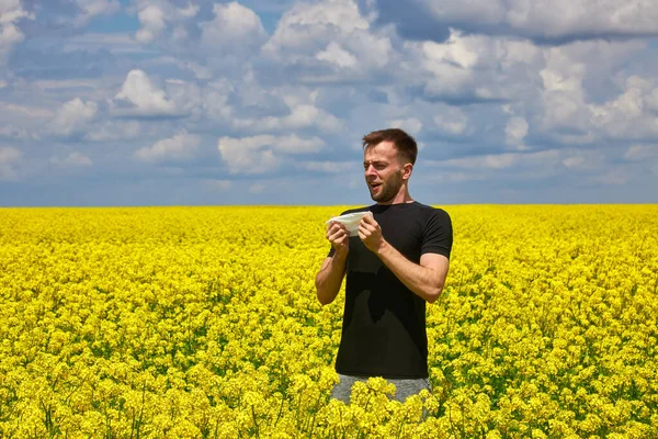 Young man in yellow canola field blowing his nose and suffering from pollen allergy.