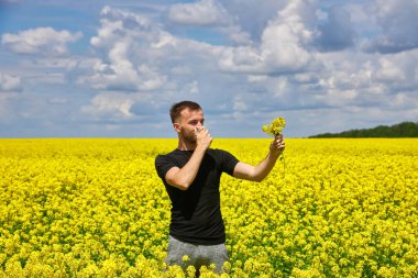 Sarı kanola tarlasındaki genç adam burnunu siliyor ve polen alerjisinden muzdarip..