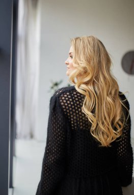 Young cute blonde posing in a studio near the window dressed in an evening black dress