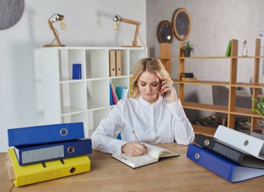 a young girl with long blond hair sits at a desk in the office, works on a computer and writes in a diary.