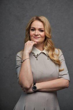 Stüdyo güzelliği, yaz elbisesi giymiş genç bir kadının portresi. Gri arka planda poz veren yazlık elbiseli sarışın kadın. Yüzüne yakın eli olan kız.