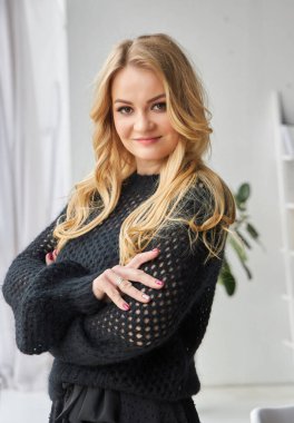 Young cute blonde posing in a studio near the window dressed in an evening black dress