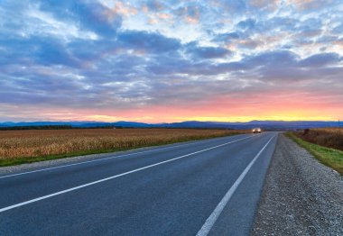 Parlak günbatımına sahip bir gece gökyüzüne karşı asfalt yolda ağaçlar hareket ediyor.