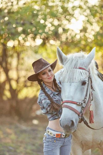 Happy beautiful young woman cowgirl in hat standing and hugging her horse