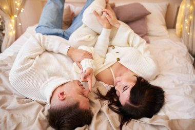 Yeni yıldan önce evde çelenklerle vakit geçiren tutkulu romantik çift manzarası. Seksi genç kadın ve yakışıklı adam yatakta kazaklarla uzanıp sarılıyorlar. Şefkatli çift birlikte yatıyor.