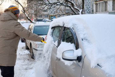 Kışın ortasında, bir adam dikkatli bir şekilde arabasından kar temizler. Güvenli sürüş koşullarını sağlamak için kar temizleme ihtiyacını yansıtan tipik bir sahne..