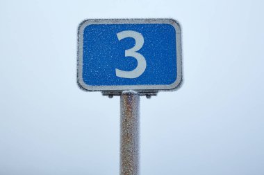 Yol kenarında buzla kaplı bir yol tabelası var. Etrafı sisli bir kış manzarasıyla çevrili..