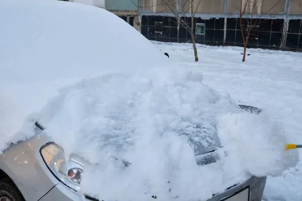 A man clearing snow from a car using a brush on a cold winter day, showcasing the routine task of snow removal