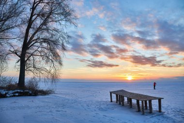 Sakin, donmuş bir gölle nefes kesici bir kış sahnesi. Güneş batarken, gökyüzü canlı renklerden oluşan bir başyapıta dönüşür. Buzlu arazinin üzerine sıcak, turuncu bir parıltı ve büyüleyici bir ahşap iskele bırakır..