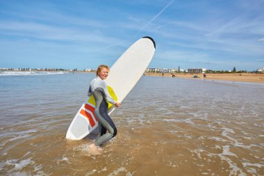 Genç bir kadın sörfçü sörf tahtası ile Essaouira 'nın okyanus kıyısında poz verir, sörf kültürünün ve sahil cazibesinin zarif bir birleşimini somutlaştırır..