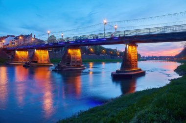 Gece şehrinin güzel manzarası, önplanda bir nehir ve yaya köprüsü var, nehirde gece şehrin ışıkları sergileniyor.
